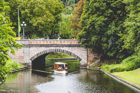 Sunlight through the trees in the park and the tourist boat sailing under the bridge through the channel. Riga, Latviaのeditorial素材