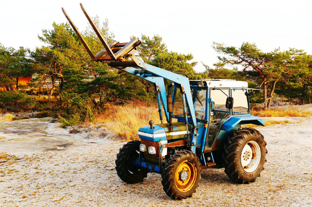 Old blue tractor. Raised excavator bucket. Large black tires. Standing on white gravel. Around the twisted pine trees. Norwegian winter. Telemark region of Ostlandet. Eastern Norway.の写真素材