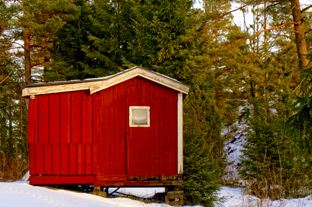 Red wooden house on the tool. Around the green conifers. Covered with snow. The sun low on the horizon. Coniferous forest around. Kragero, Telemark municipality. Region of southeastern Norway. Skagerrak coast.のeditorial素材