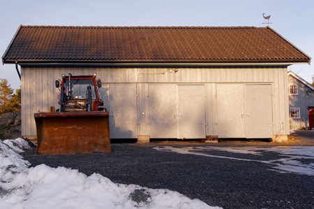 Tractor with plow snow. Old tractor parked in front of a large garage. On the black gravel. Winter scenery, snow on the rocks. Kragero, Telemark municipality. Region of southeastern Norway. Skagerrak coast.のeditorial素材