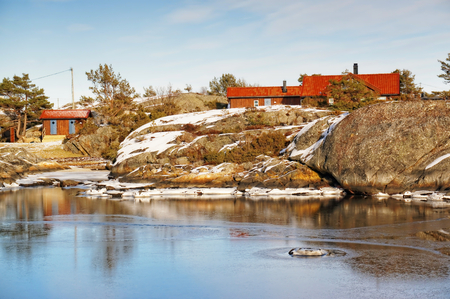 Rd summer cottage hidden on the rocks. Near bay fjord covered in ice in the winter. Wavy rocks covered with snow. In the background of the North Sea. Visible signs of outflow of the sea. Blue sky. Winter in Aust-Agder municipality. Region of southeastern のeditorial素材
