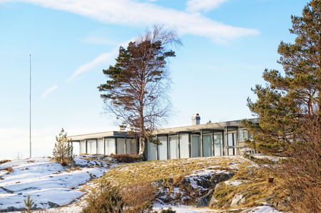 Grey long and flat guest cottage, with large windows. Hidden behind a rock and trees on the coast. Blue sky. Wavy rocks covered with snow. Winter in Aust-Agder municipality. Region of southeastern Norway. North Sea Coast. Skagerrak coastline. Norwegian laのeditorial素材
