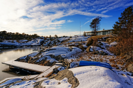 Long wooden pier, covered with snow, over fjord. In the background of long and flat cottage, with large windows. Hidden behind a rock and trees on the coast. Next to you empty flag. Blue sky. Birch and pine among the rocks.のeditorial素材