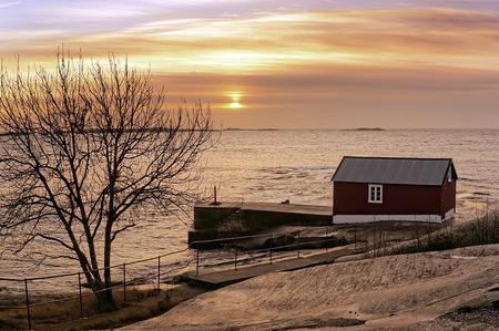 Winter view of the sunrise over the fjord. Rocks lit by sunlight, the remnants of snow. Red wooden building on the shore fishing.の写真素材