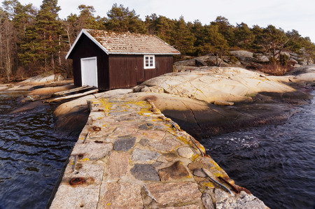 Old brown wooden boat building. Rocks fjord in early spring. Region of southern Norway. Evergreen trees, pines, spruces. Trees and shrubs without leaves. Sand and rocks.のeditorial素材