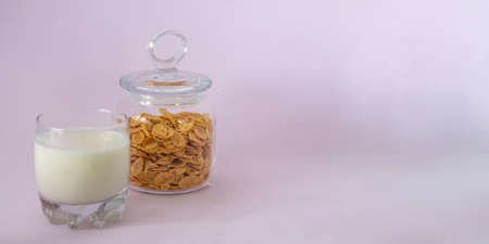 a glass of milk and a glass jar with cornflakes on a solid pink background. bannerの写真素材