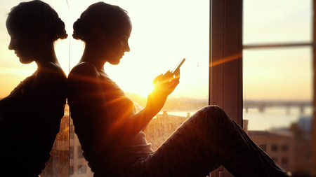 beautiful young woman sitting on window uses smart phone on background of the sunset and cityの写真素材
