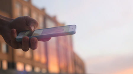 Mans hand touch screen smart phone on blurred architectural building during sunset time.の写真素材