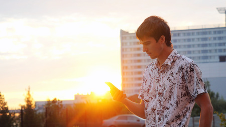 Young handsome man using smartphone outdoors at beautiful sunset time. Businessman walking on the city background through the sun with lense flare effectsの写真素材