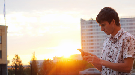 Young man using smartphone outdoors at beautiful sunset time. Businessman walking on the city background through the sun with lense flare effectsの写真素材