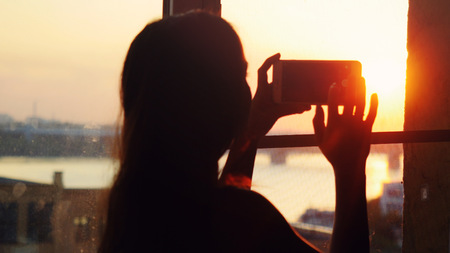 Young pretty woman taking photos of city with blurred architectures on mobile smart phone at home window during sunset with beautiful lense flare effects.の写真素材