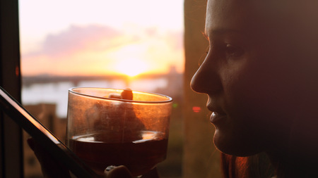 Young woman uses the phone, standing at the window drinks coffee on the background of a beautiful sunset with sun flareの写真素材