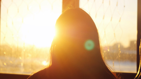 Woman looking standing near the window with view on sunset in city and lense flare effectsの写真素材