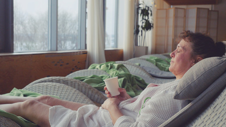 Mature woman in aged wears white dressing gown in spa centre lying on the sofa drinks tea and looks in the window with quiet view of the river and city urban backgroundの写真素材