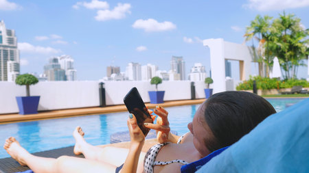 Young woman using smartphone sends sms on sunbed by pool on urban architecture background