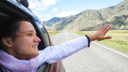 Smiling young brunette woman in a car playing with wind and driving past the beautiful mountainsの写真素材