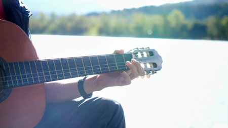 Close up of hand guy plays on a guitar sitting by mountain river on sunny day with lens flare effectsの写真素材