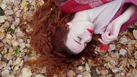 Beautiful young woman with red hair lies over dry leaves holds a mountain ash in autumn forestの写真素材