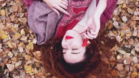 Attractive young woman with red hair lies over dry leaves holds a mountain ash in her hand in autumn forestの写真素材