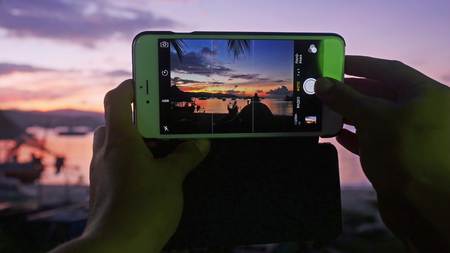 Close up of hands of woman taking photo with mobile phone of sunset while sitting in the beach.の写真素材