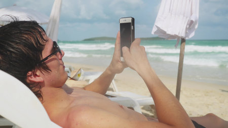 Young man using smartphone while lying on sunbed on tropical beach with turquoise sea background.の写真素材