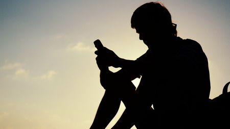 Silhouette of young man using a cellphone on the rock at the sea during beautiful sunset.の写真素材