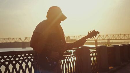 Russia, Novosibirsk, 27 july, 2017. Young caucasian man standing outdoors in promenade and playing guitar at amazing sunset with lens flare effects.のeditorial素材