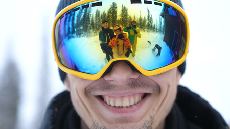 Close up of portrait of smiling man in the blue skiing glasses in deep beautiful forest and cheerful friends waving their hands and are reflected in his glasses.の写真素材