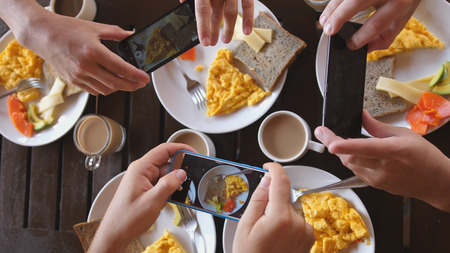 Top view of a group of friends having breakfast in a cafe at wooden table, taking picture of omelette, toasts and coffee with mobile phone.の写真素材