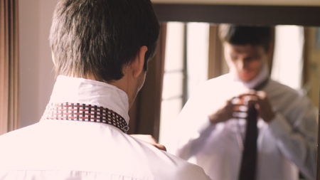 Man in white shirt stands by the mirror tying a tieの写真素材