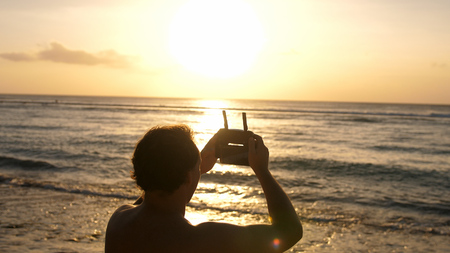 Man operating a drone with remote control. Quadro copter drone flying over sunset oceanの写真素材
