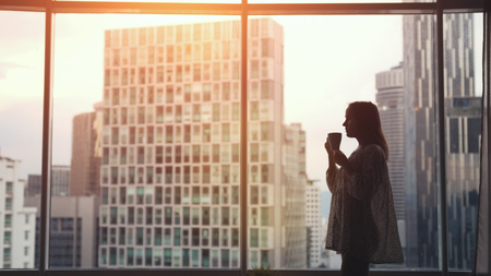 Beautiful woman drinking coffee by the window with amazing cityscape view during sunsetの写真素材
