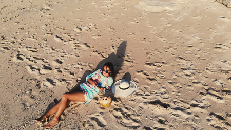 Top view of young brunette woman in sunglasses lying on tropical paradise beach and uses mobile phone during beautiful sunset. Travel conceptの写真素材