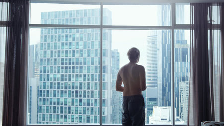View from behind of young man in shorts looking out at view of the city. View from the window shows...