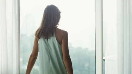 Young brown-haired woman looking through window to the city view.の写真素材