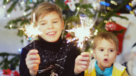 Happy boys burn sparklers among bokeh lights and garlands on a Christmas tree