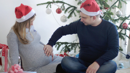 Happy young family. Husband touches belly his pregnant wife near beautiful decorated Christmas tree.の写真素材