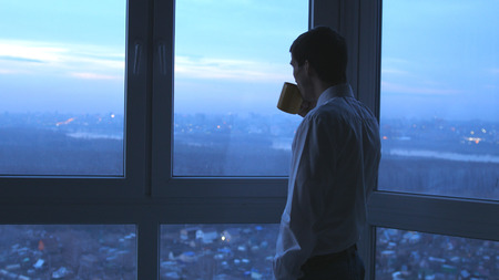 Young businessman drinking tea and admire view from window in the evening in officeの写真素材