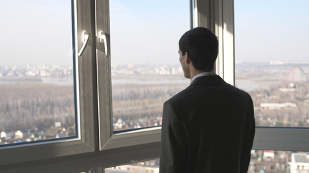 Back view of young successful businessman standing in front of windows looking into distance on the cityの写真素材