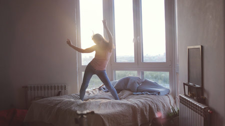 Happy young woman jumps and dances on bed in luxury apartment during beautiful sunrise with lens flare effect in the morningの写真素材