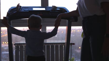 Funny little child walks on modern treadmill and mom hold his handの写真素材