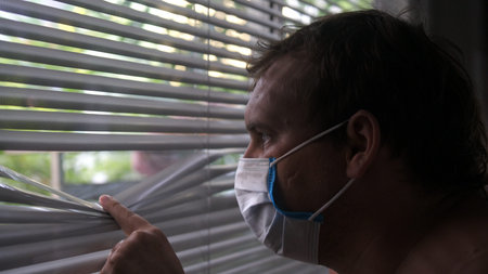 Sad young man in protective medical mask looks over window blinds