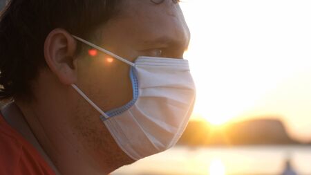 A masked young sad man sits on the beach at sunset time. Coronavirus epidemicの写真素材