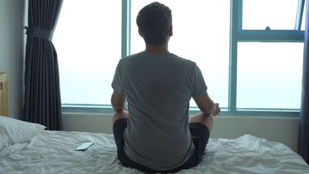 A man meditates on bed while looking out the window. Back viewの写真素材