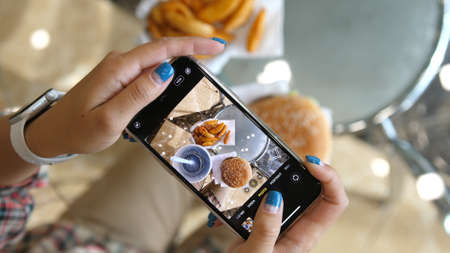 Woman using phone to take pictures of Burger, french fries and cola. Tourist girl have brunch or lunch time at fast food cafe. Top view. Novosibirsk, Russia. 2020のeditorial素材