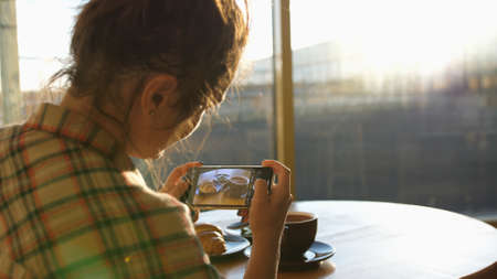 Close up of woman holds modern smartphone and taking pictures of her morning breakfast coffee and tasty croissant buns to share photos on social media resourcesの写真素材