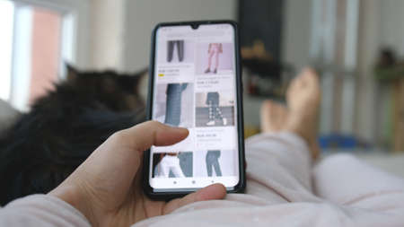 A Woman at home Lying on sofa with cat in living room looks at Goods in Online Store Clothing Using Smartphone Buys in Internet Shop.の写真素材