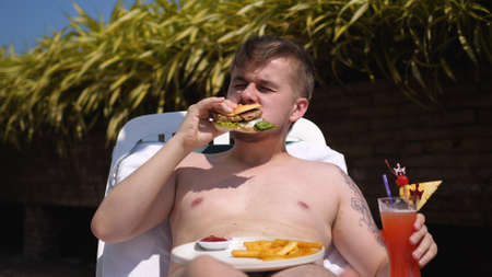 Bearded man is eating a hamburger lying on a lounger with a cocktail. Junk food. unhealthy eat habits.の写真素材