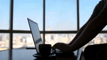 A concentrated young businessman is working on a laptop computer using his notebook sitting by the window with urban view and drinks coffeeの写真素材