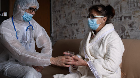 Kind doctor in protective suit giving Giving A Glass Of Water to senior woman wearing medical mask, medical careの写真素材